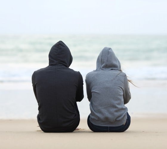 To ungdommar med hettegenser sett bakfrå som sitt på ei strand